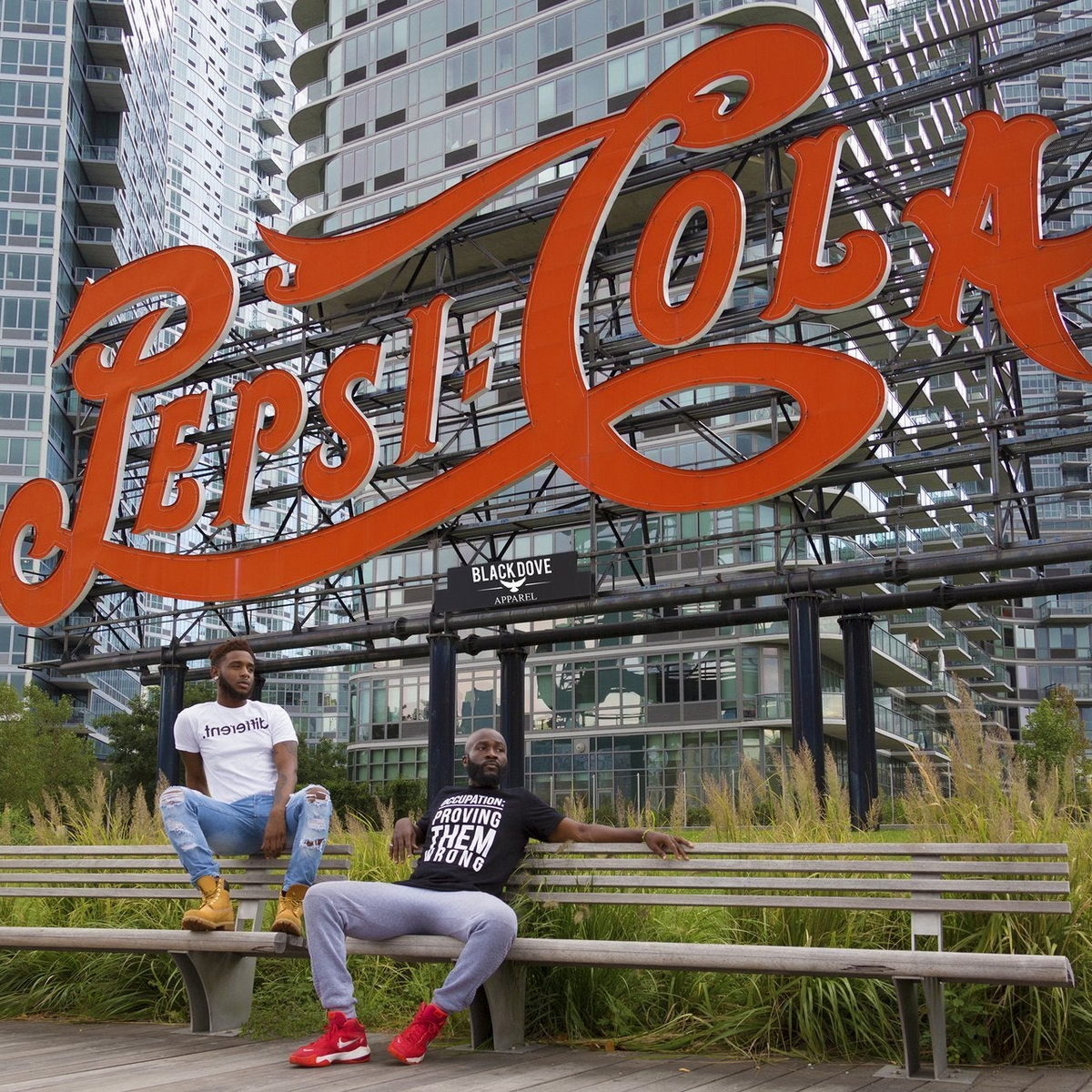 Two people sitting on a bench in front of a large Pepsi Cola sign with urban buildings in the background.