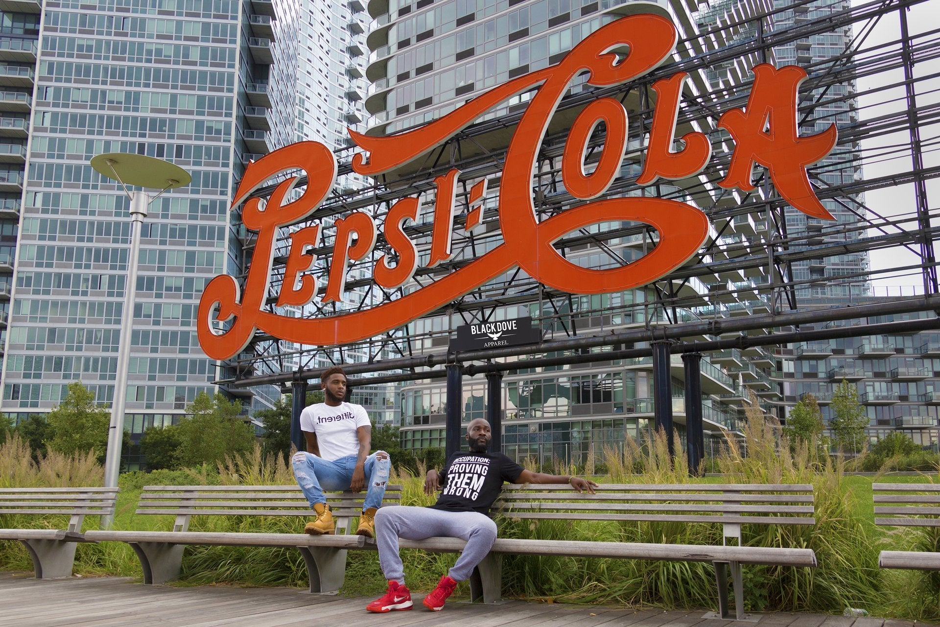 Two people sitting on a bench in front of a large Pepsi Cola sign with urban buildings in the background.