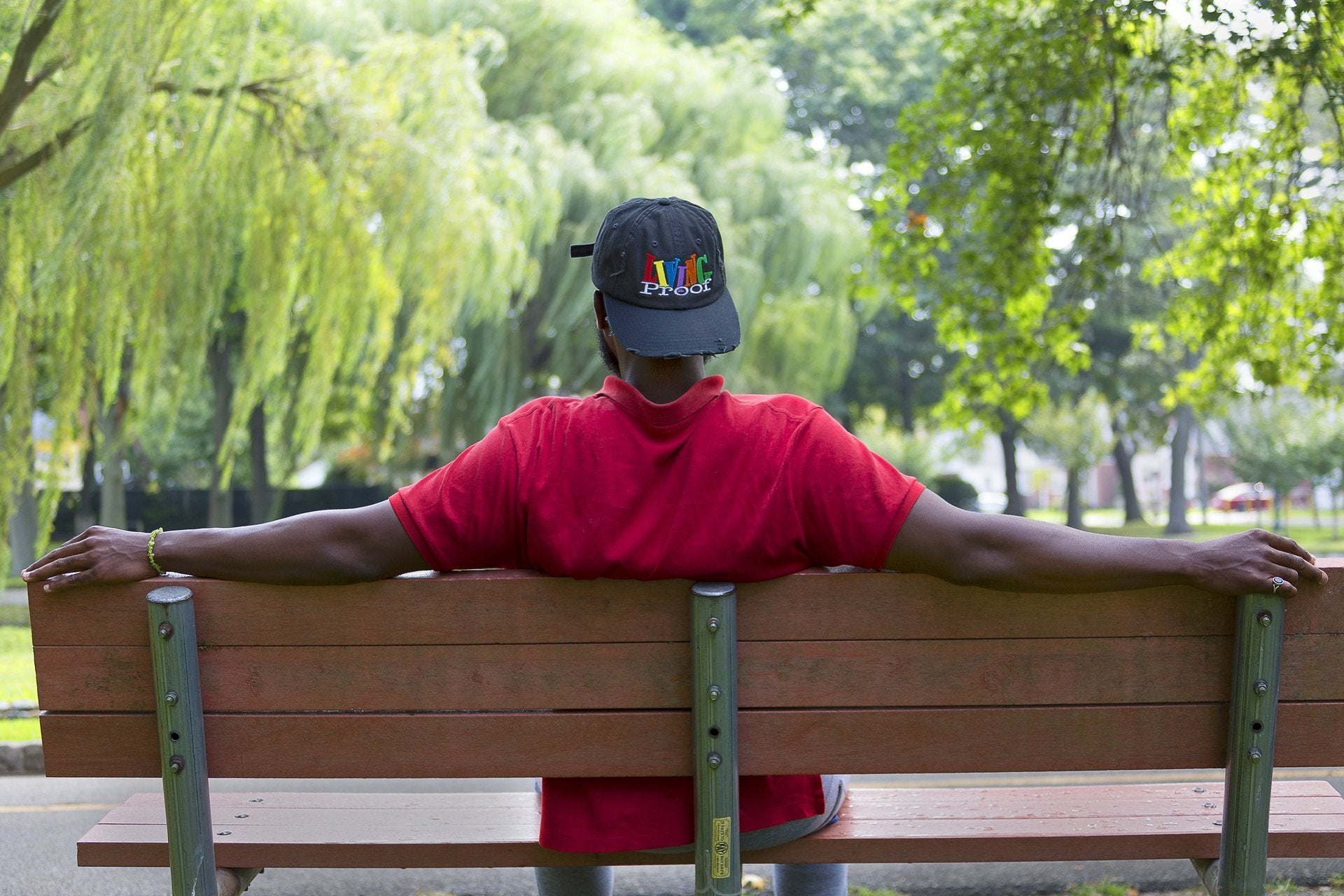 Person wearing a colorful cap and red shirt sitting on a bench in a park.